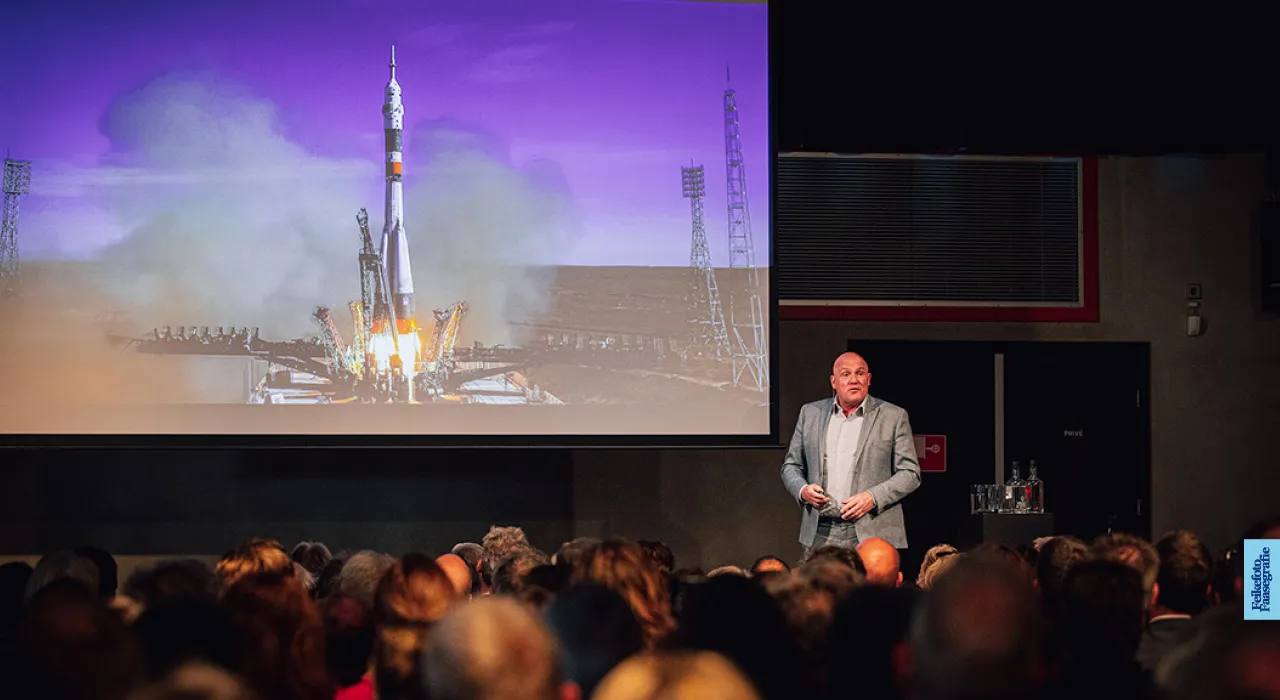 Astronaut André Kuipers geeft een presentatie op een podium voor een volle zaal. Op het grote scherm achter hem is een spectaculaire lancering van een Sojoez-raket te zien met veel rook en vuur bij het opstijgen.