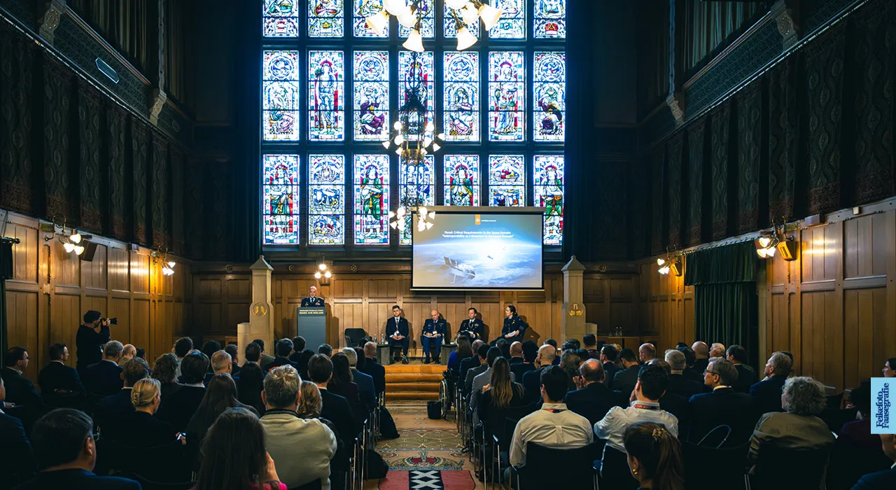 Een paneldiscussie over ruimtevaart en defensie op het podium van de Effectenbeurszaal in de Beurs van Berlage te Amsterdam. Op de achtergrond is het iconische, verlichte glas-in-loodraam te zien en de zaal zit vol met publiek.
