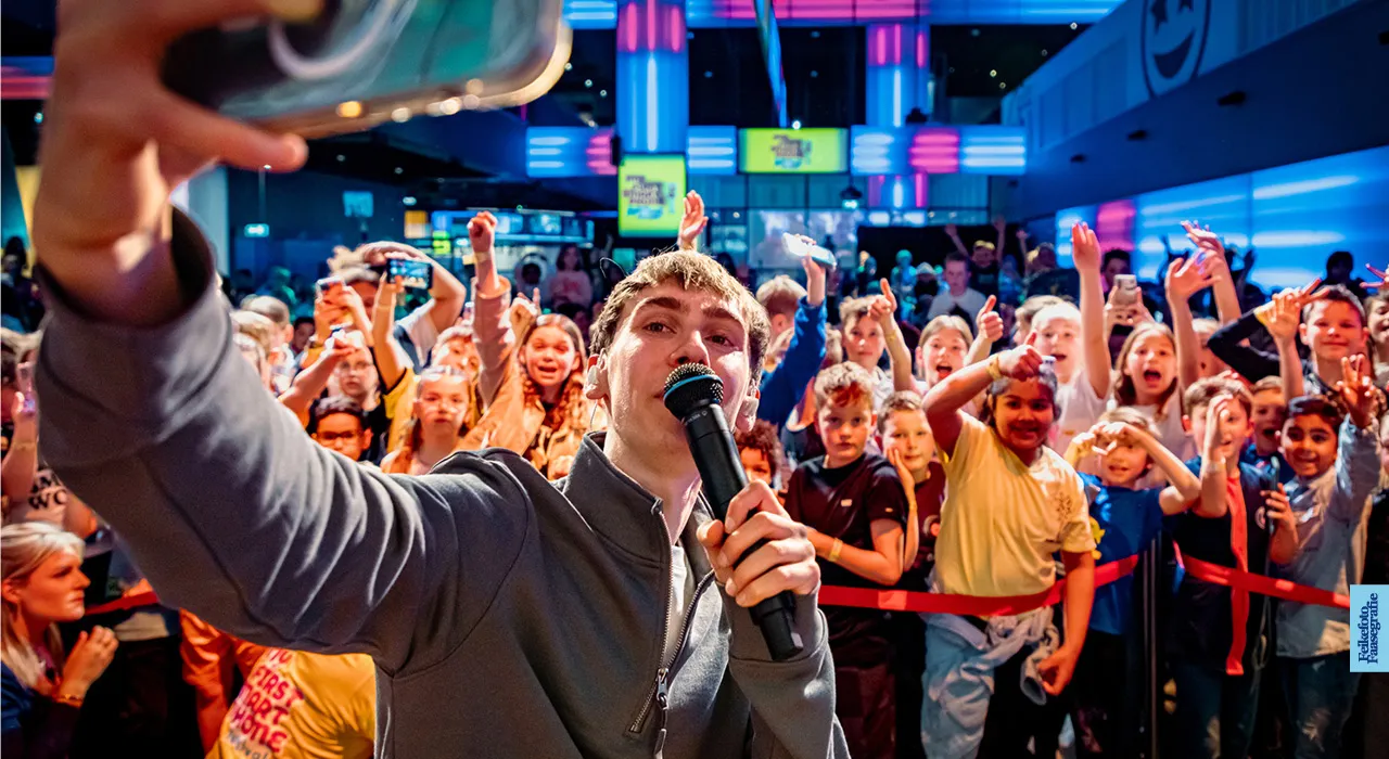 YouTuber DutchTuber maakt een selfie met een microfoon op het podium tijdens het My First Smartphone Festival in Beeld & Geluid, met een enthousiast publiek van kinderen achter hem.