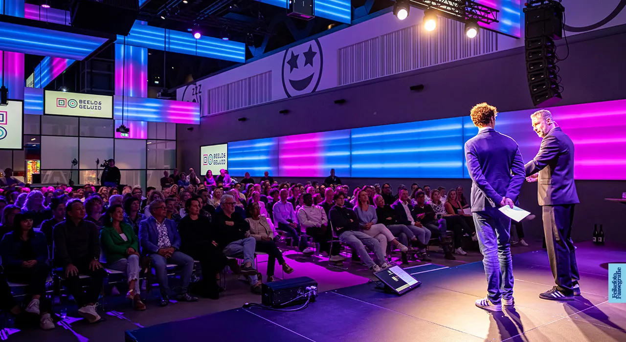 Twee mannen op een verlicht podium presenteren voor een grote zaal met zittend publiek tijdens een evenement in Beeld & Geluid, met opvallende blauwe en paarse neonverlichting aan de wanden.