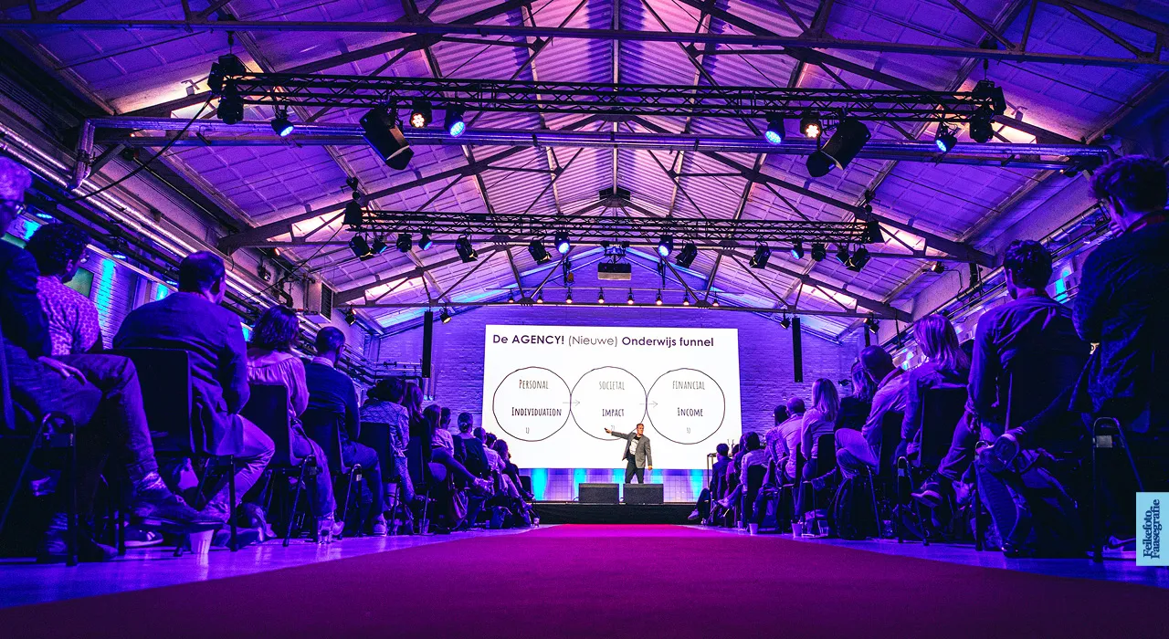 Een spreker geeft een presentatie over de nieuwe onderwijs funnel in een grote industriële evenementenhal met paarse belichting. Op het grote scherm is een diagram te zien met 'Personal Individuation', 'Societal Impact' en 'Financial Income'. Het publiek kijkt vanaf weerszijden van een rode loper toe.