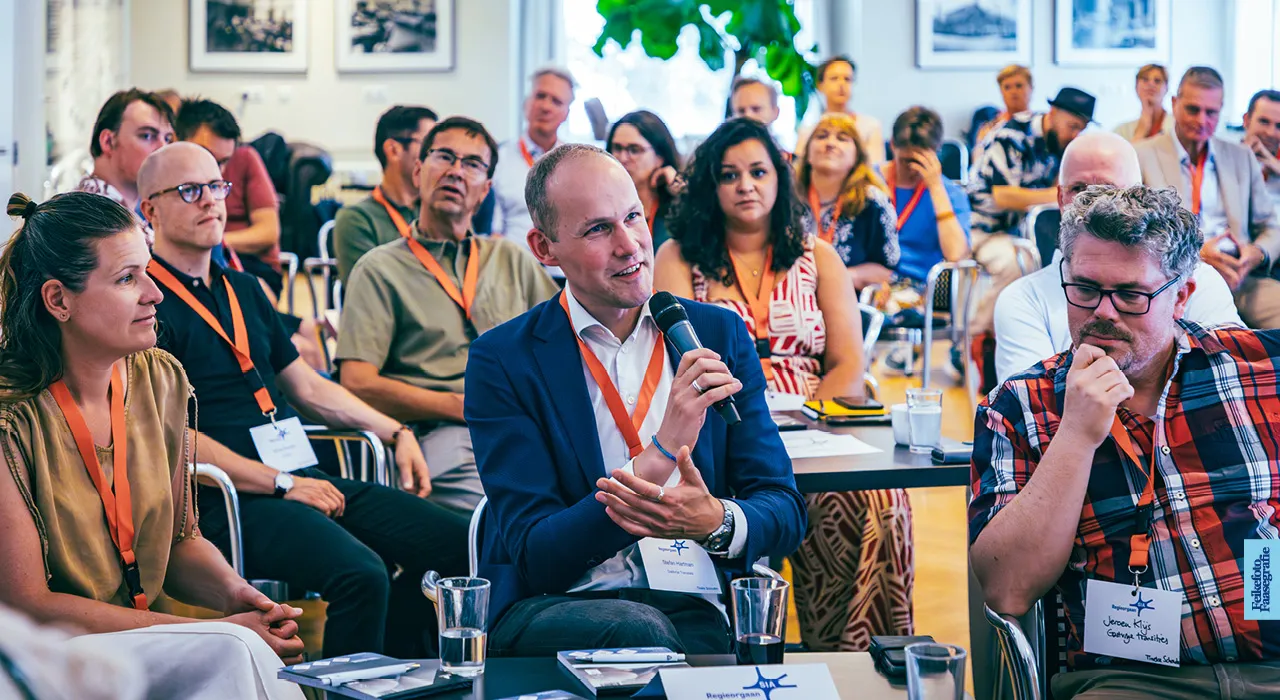 Stefan Hartman stelt een vraag met een microfoon tijdens een interactieve bijeenkomst of congres van Regieorgaan SIA. Naast hem luisteren andere deelnemers, waaronder Jeroen Klijns, aandachtig mee in een lichte vergaderruimte.