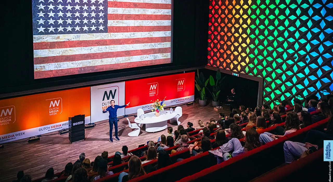 Amerika-correspondent en journalist Michiel Vos geeft een enthousiaste lezing op het podium van Dutch Media Week in Beeld & Geluid Hilversum. Op het grote bioscoopscherm achter hem staat een geverfde Amerikaanse vlag. De zaal zit vol met publiek.