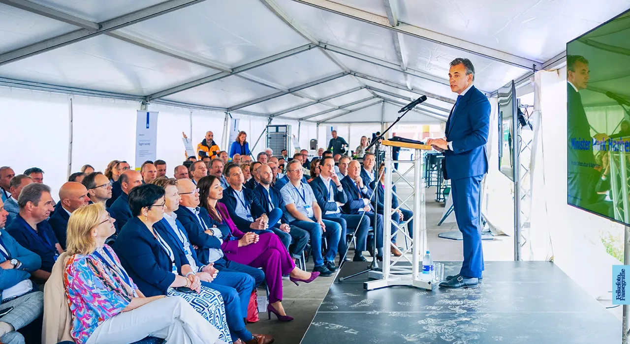 Minister Mark Harbers van Infrastructuur en Waterstaat spreekt tijdens een bijeenkomst voor het project 'Verruiming Twentekanaal'. Hij staat op een podium in een evenemententent voor een zittend publiek van betrokkenen en genodigden.