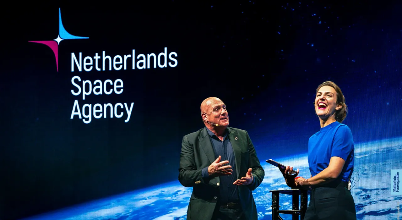 Een man en een vrouw presenteren lachend op een podium tijdens een evenement van de Netherlands Space Agency. Op de achtergrond is een groot scherm te zien met het logo van de organisatie en een afbeelding van de aarde vanuit de ruimte.