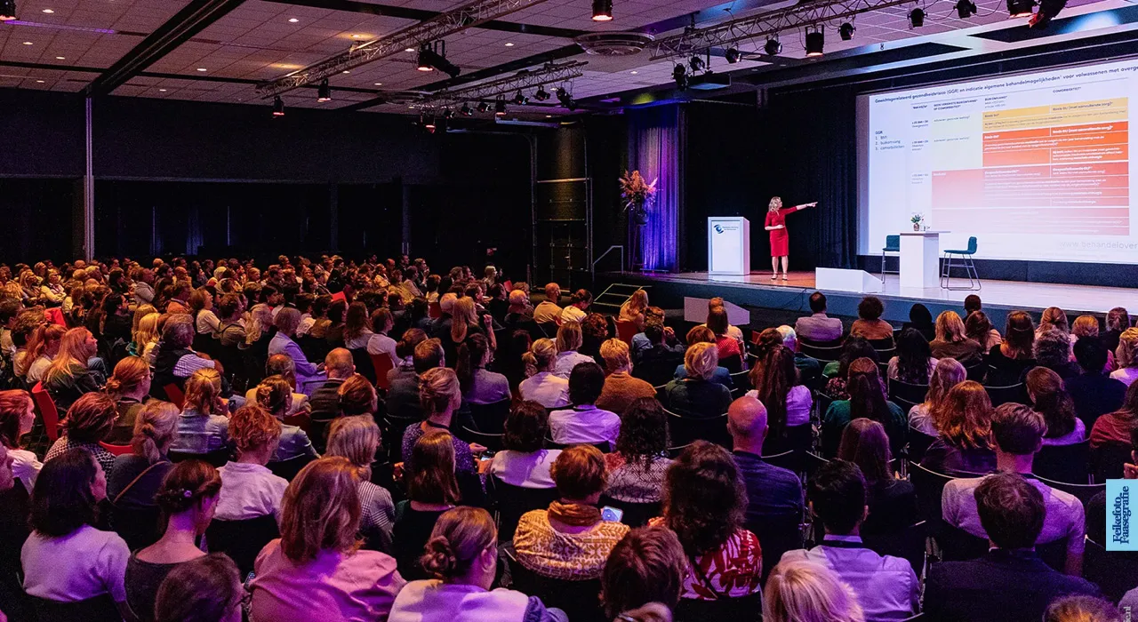 Een vrouwelijke spreker in een rode jurk geeft een presentatie over gewichtsgerelateerd gezondheidsrisico (GGR) namens het Partnerschap Overgewicht Nederland (PON) tijdens een evenement voor de vereniging van doktersassistenten (NVDA). De zaal zit vol met aandachtige luisteraars.