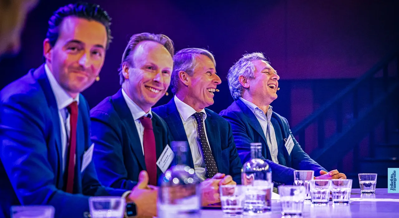 Vier mannen in pak, waaronder ondernemer Ali Niknam, lachen tijdens een paneldiscussie op een congres in Theater Spant te Bussum, zittend aan een tafel onder paarse evenementenverlichting.