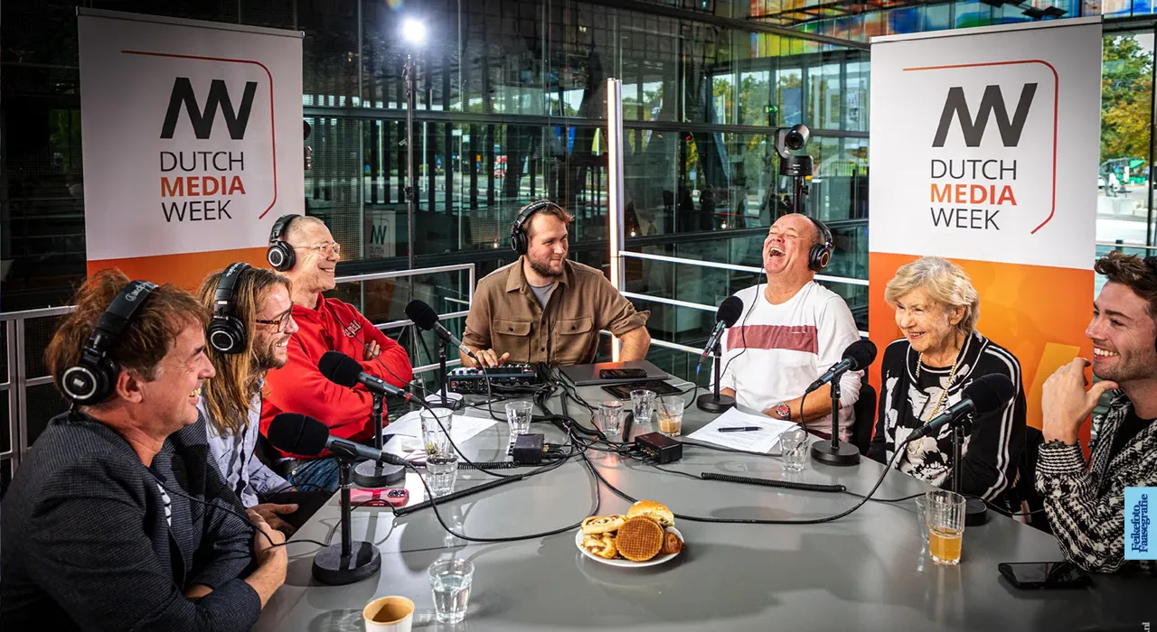 Een vrolijke live podcast-uitzending aan een ronde studietafel tijdens Dutch Media Week, met een breed lachende Paul de Leeuw, zijn moeder, Hans Kesting en Giel Beelen met koptelefoons op en microfoons voor zich.