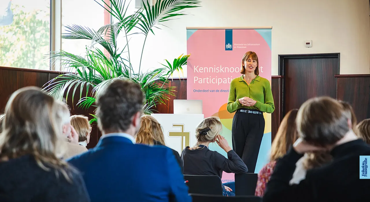 Een vrouw in een groene top geeft een presentatie voor een zittend publiek in Villa Jongerius te Utrecht, met op de achtergrond een banner van het Kennisknooppunt Participatie van het Ministerie van Infrastructuur en Waterstaat.