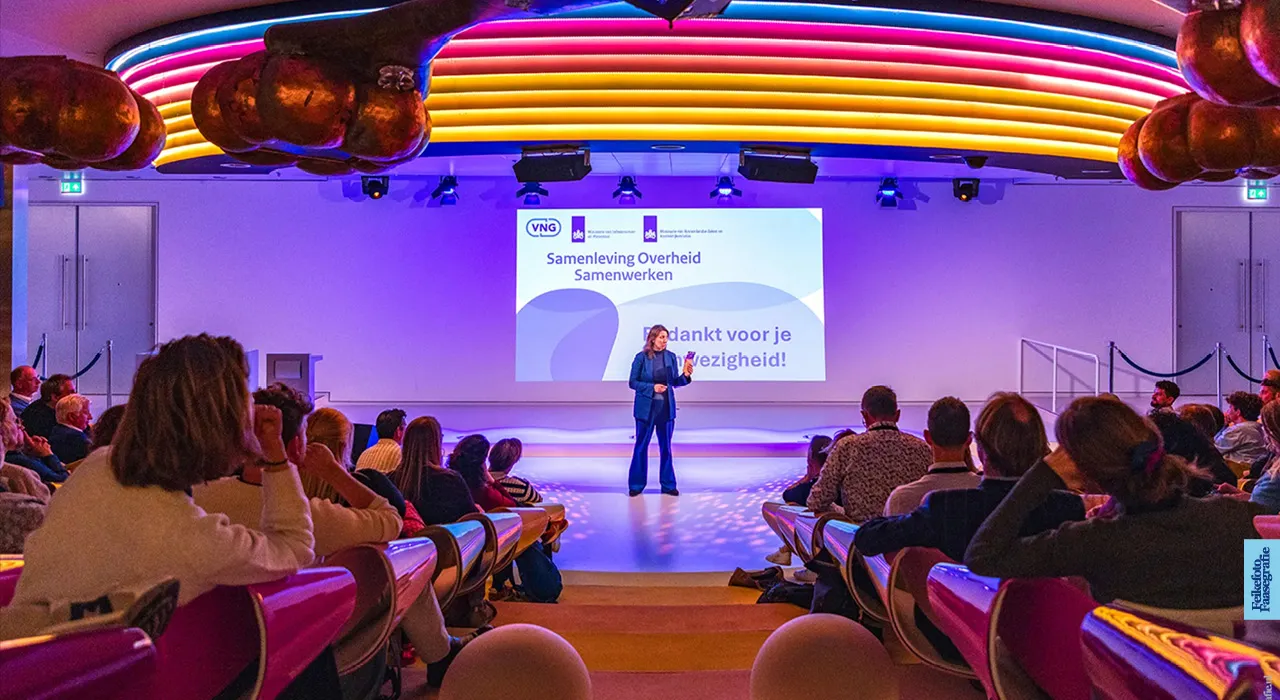 Een vrouwelijke spreker sluit het nationale S.O.S. (Samenleving Overheid Samenwerken) congres af op het podium van het Mediaplaza in de Jaarbeurs Utrecht. Het evenement is georganiseerd door de VNG en het Ministerie van IenW. De zaal heeft een opvallend, verlicht regenboogplafond.