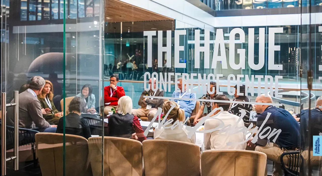 Een groep professionals in een cirkel tijdens een vergadering of breakout-sessie, gezien door een glazen wand met het logo van The Hague Conference Centre New Babylon.