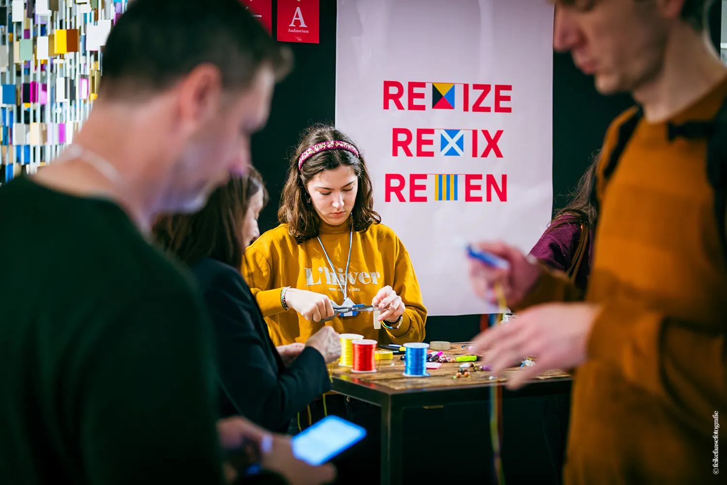 Een vrouw in een mosterdgele trui werkt geconcentreerd met een schaar en kleurrijke garen aan een tafel tijdens een creatieve workshop. Op de achtergrond hangt een poster met de tekst 'RESIZE REMIX RETHINK'. Andere deelnemers zijn onscherp op de voorgrond zichtbaar in een dynamische evenementensetting.